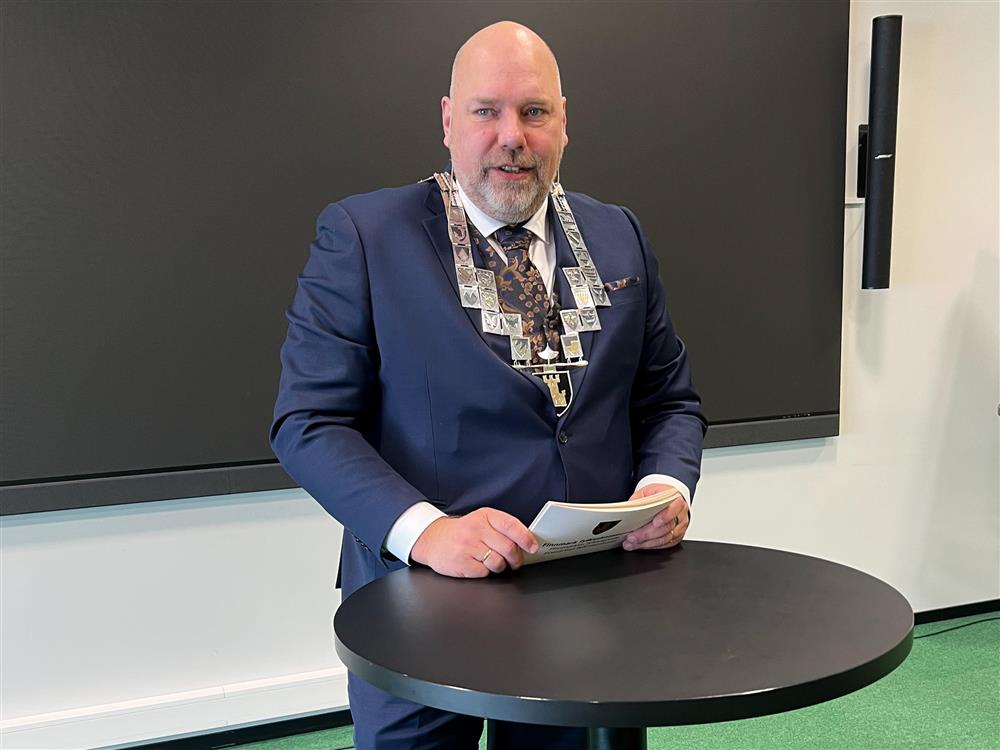 En mann i dress og ordførerkjede står ved et bord. I hendene holder han talekort. Han ser til venstre i bildet. - Klikk for stort bilde