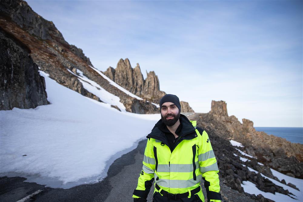 En mann i varselgul jakke smiler mot kamera. Bildet er tatt på en landvei på vinterstid. - Klikk for stort bilde