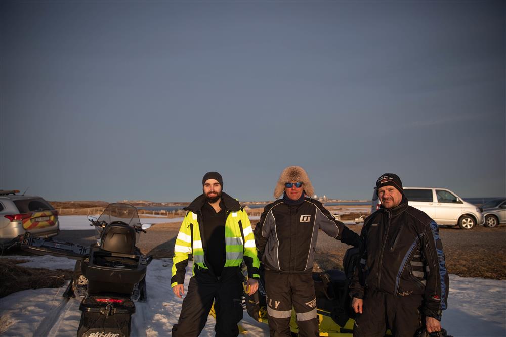 Tre menn i solnedgangen smiler til kamera. De er godt kledd, bildet er tatt i skiftet mellom vinter og vår. - Klikk for stort bilde
