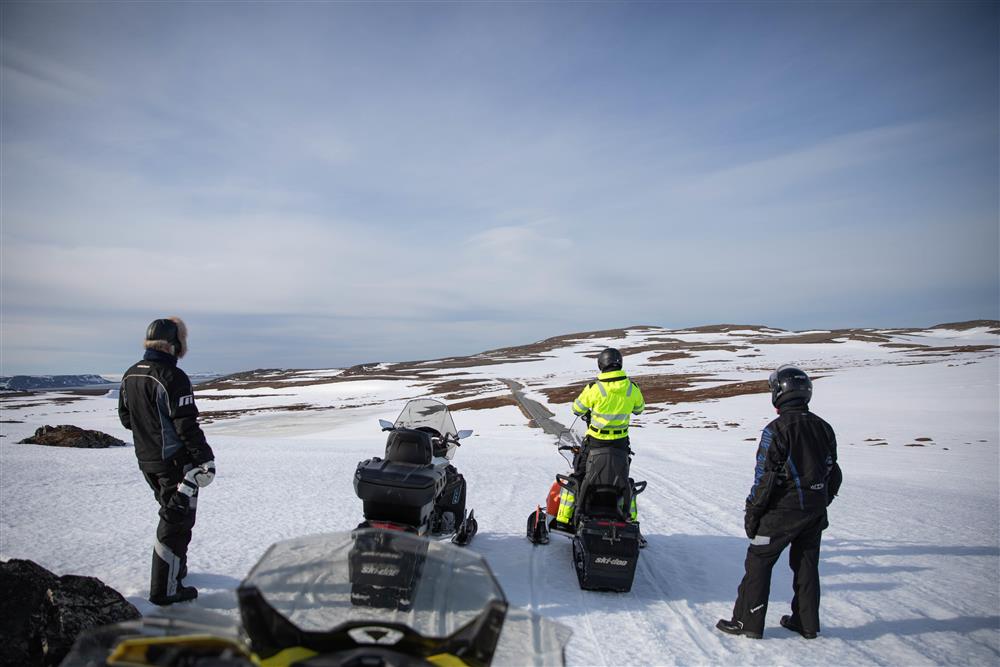 Flere personer står ved snøscooteren sin i et arktisk landskap på vinterstid. De skuer over en landvei. - Klikk for stort bilde